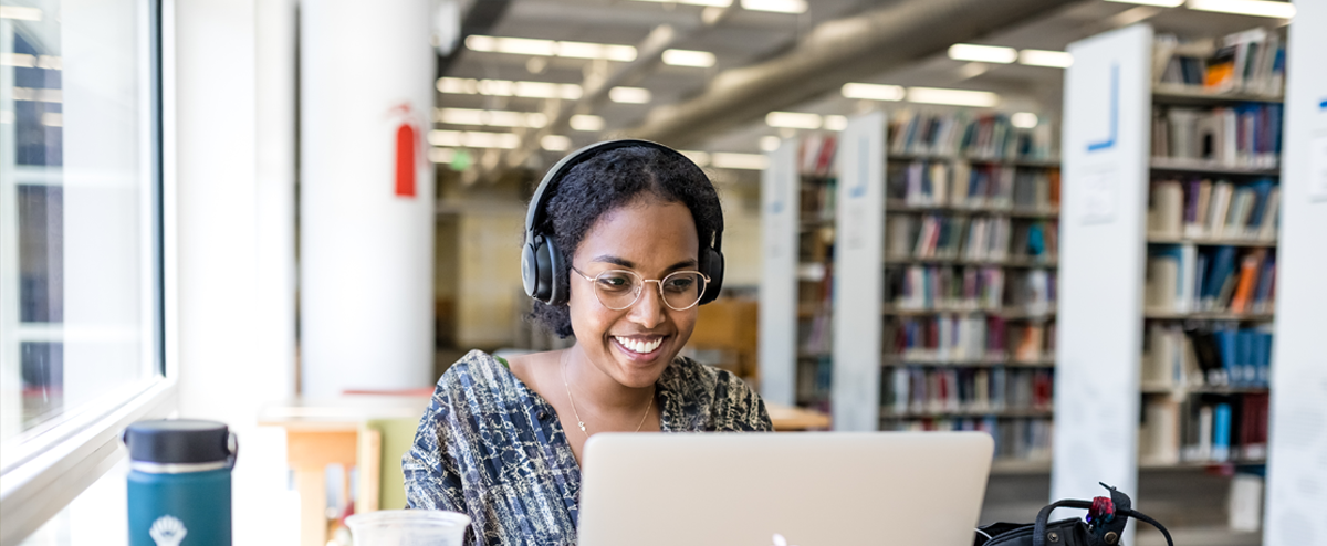 Image of a student sitting in front of a computer with headphones on in the library, smiling.